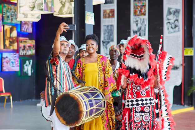 Photo 2: The Honourable Michelle Benjamin, Minister of Culture and Community Development poses for a “selfie” with local musicians and Carnival characters during the production for Trinidad and Tobago’s CARIFESTA XV promotional video at the Queen’s Park Savanah, Port of Spain.